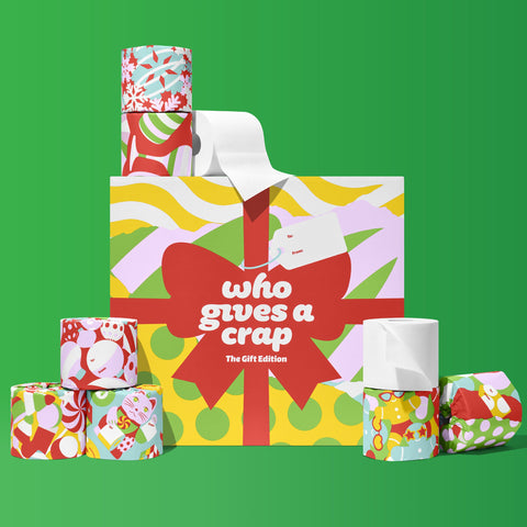 A 48-roll box of Who Gives A Crap The Gift Edition toilet paper in red, lime and pink gift-wrap design, stacked with individually wrapped rolls on a green background.