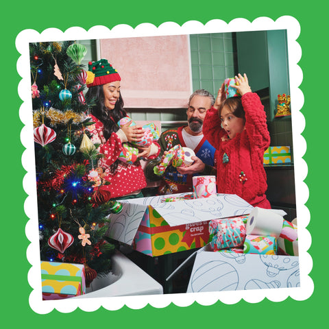 A family in festive jumpers unwraps Gift Edition toilet paper next to a decorated Christmas tree in a green-tiled bathroom.